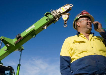 Rigger apprentice in hi-vis shirt and hard hat holding a radio beside a green crane boom, Artisan Training Institute